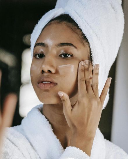 Young black female  in white robe and towel on head  applying moisturizing cream on face while standing in bathroom